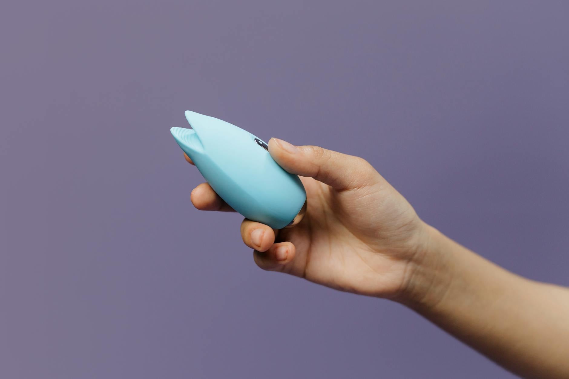 A blue silicone sex toy held against a solid purple background, symbolizing touch and clitoral stimulation.